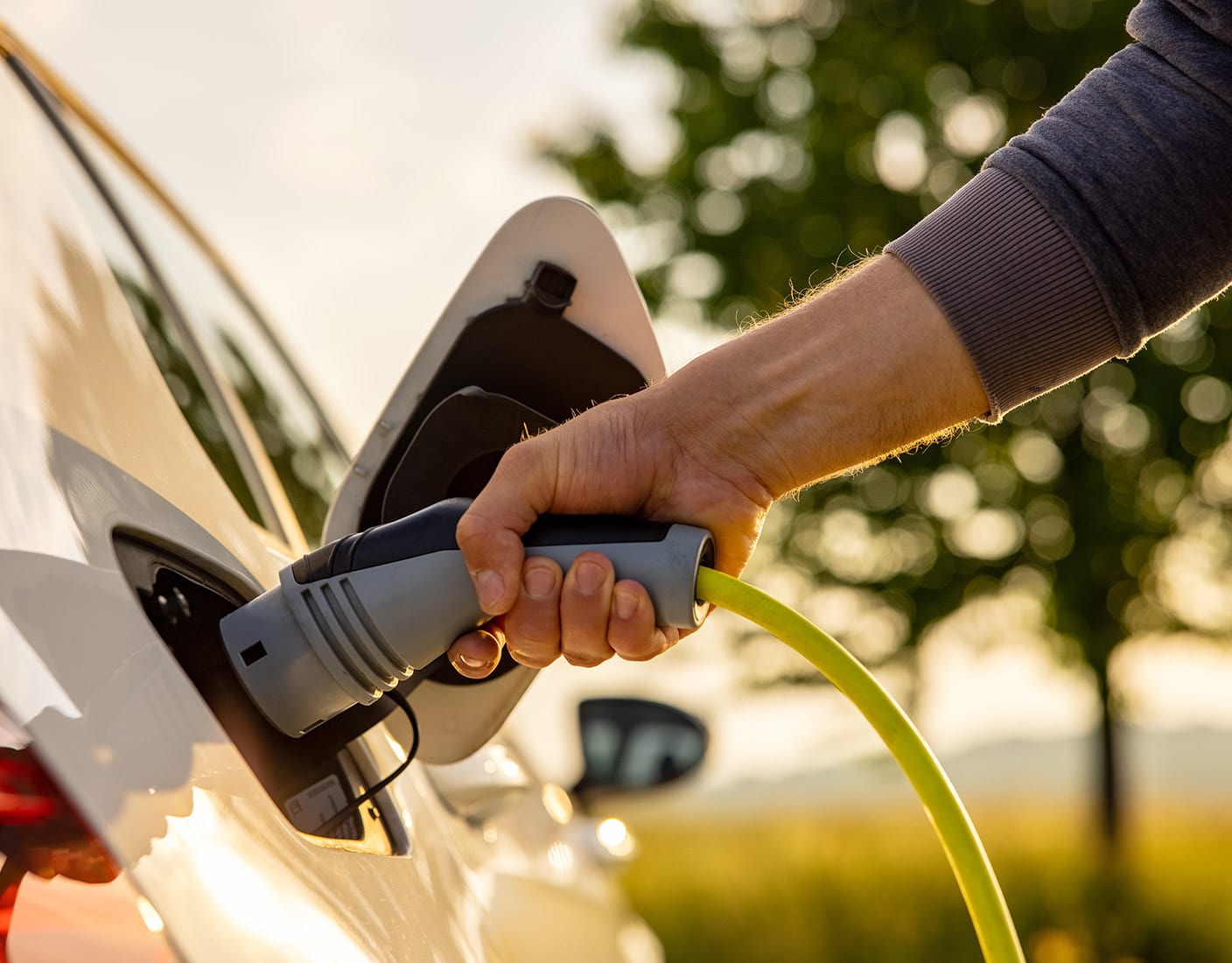 A hand plugs a charging cable into a white electric car, with a blurred outdoor background.
